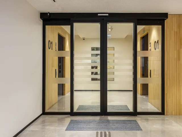 Automatic fire-rated sliding glass doors with black frames leading to a hallway with numbered doors.
