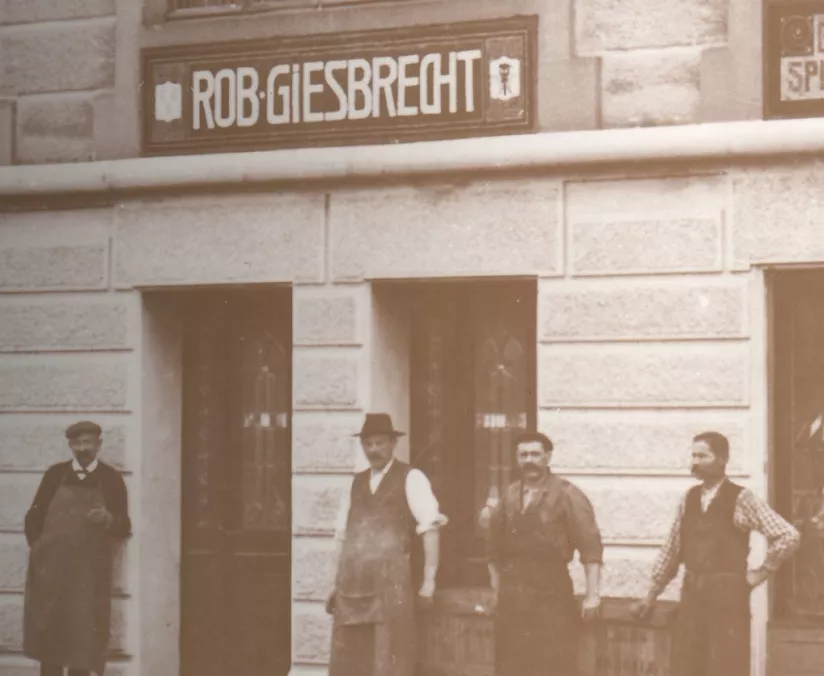 Giesbrecht History 1884 - Junkergasse, Bern Historical Vetrotech image showing a storefront with period signage and men, highlighting its long history.